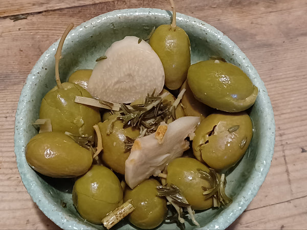 A small speckled light blue bowl filled with green olives, garlic cloves, and herbs, set on a rustic wooden table. A 'Galaxy A32 5G' watermark is visible in the bottom-left corner.